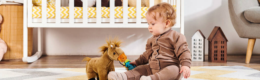 милый маленький мальчик играет с игрушечной лошадью рядом с кроваткой на полу в детской комнате, горизонтальный баннер - Фото, изображение