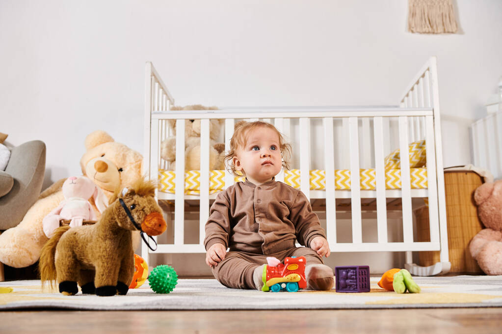 маленький невинный мальчик, сидящий на полу рядом с кроваткой и мягкими игрушками в уютной детской комнате, блаженное детство - Фото, изображение