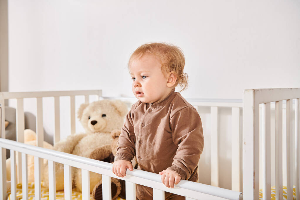 幸せな子供時代,愛らしい幼児の少年は自宅で居心地の良い保育室で柔らかいおもちゃでベビーベッドに立っています - 写真・画像
