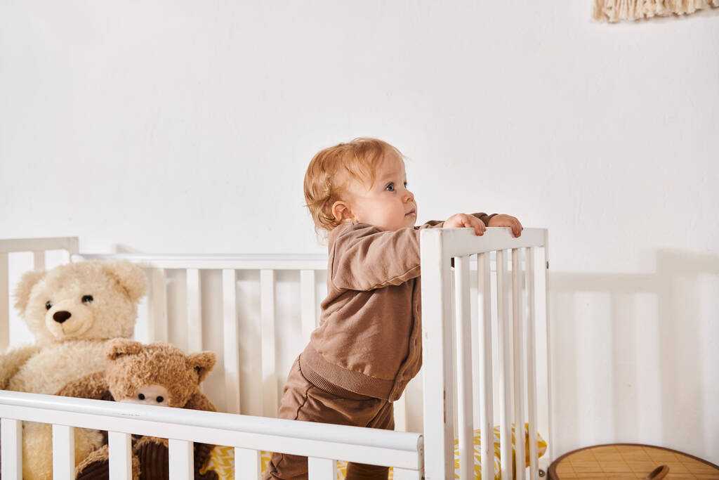 ciekawy chłopiec stojący w łóżeczku z miękkimi zabawkami w przytulnym pokoju dziecięcym w domu, błogie dzieciństwo - Zdjęcie, obraz