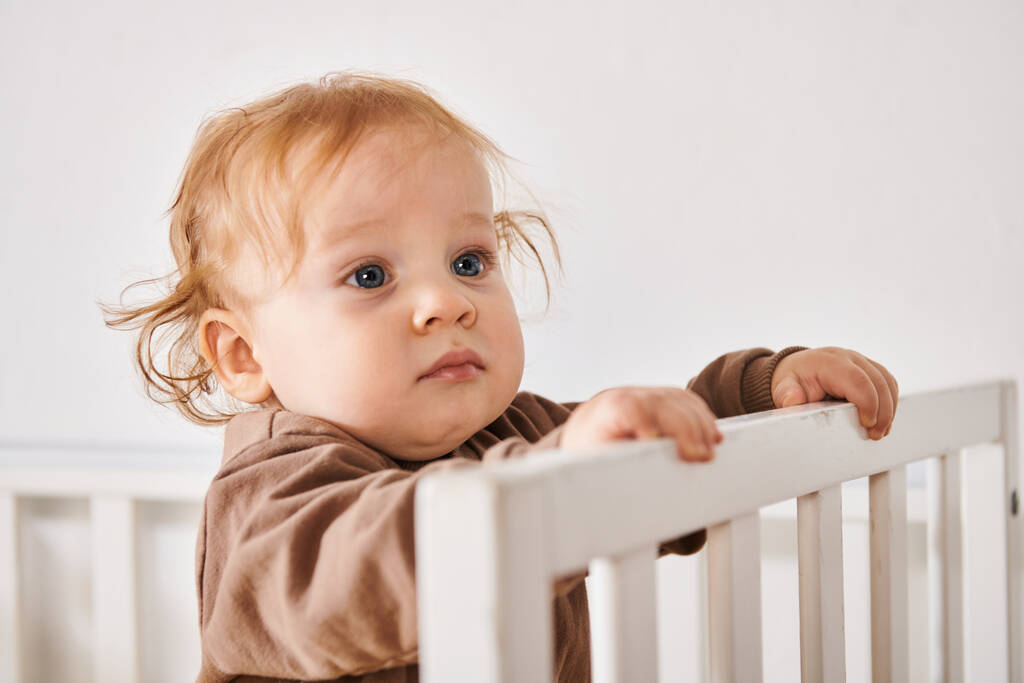 Porträt eines entzückenden Kindes, das in der Krippe im Kinderzimmer steht und wegsieht, glückliches Baby - Foto, Bild