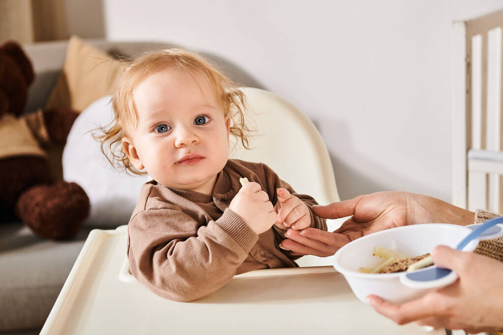 восхитительный мальчик, сидящий в детском кресле и смотрящий на камеру рядом с мамой с миской завтрака - Фото, изображение