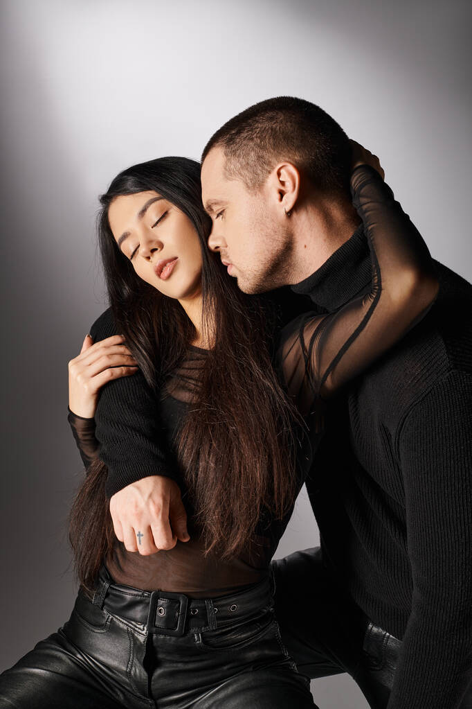 romántico interracial pareja en negro casual atuendo abrazando con cerrado ojos en gris telón de fondo - Foto, Imagen