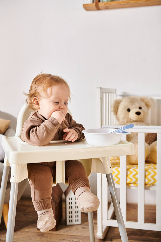 χαριτωμένο μικρό παιδί κάθεται σε καρέκλα μωρό κοντά μπολ με πρωινό στο παιδικό δωμάτιο, ευτυχισμένη παιδική ηλικία - Φωτογραφία, εικόνα