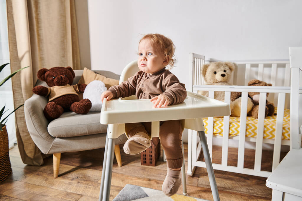 маленький хлопчик сидить у дитячому кріслі і дивиться в затишну дитячу кімнату з ліжечком і плюшевими ведмедями - Фото, зображення