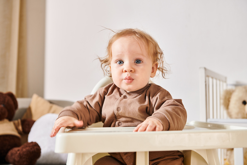 retrato de lindo niño sentado en silla de bebé y mirando a la cámara en la habitación de la guardería en casa - Foto, Imagen