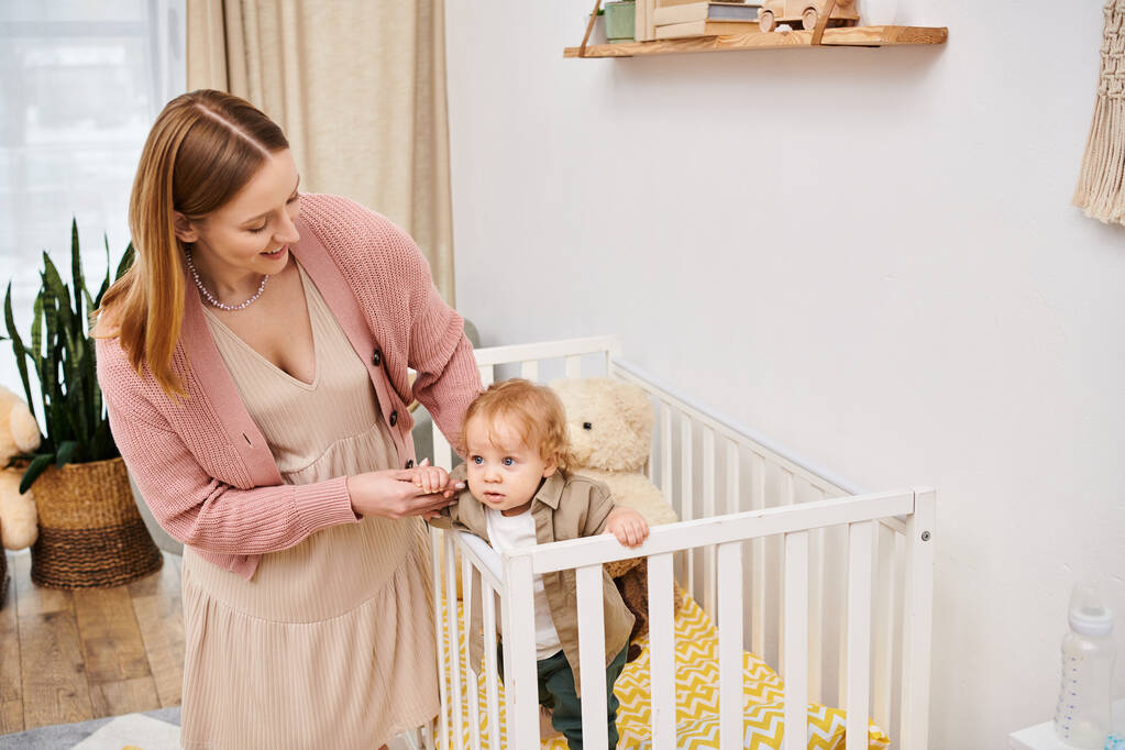 young woman supporting little boy standing in crib in nursery room at home, blissful motherhood - Photo, Image