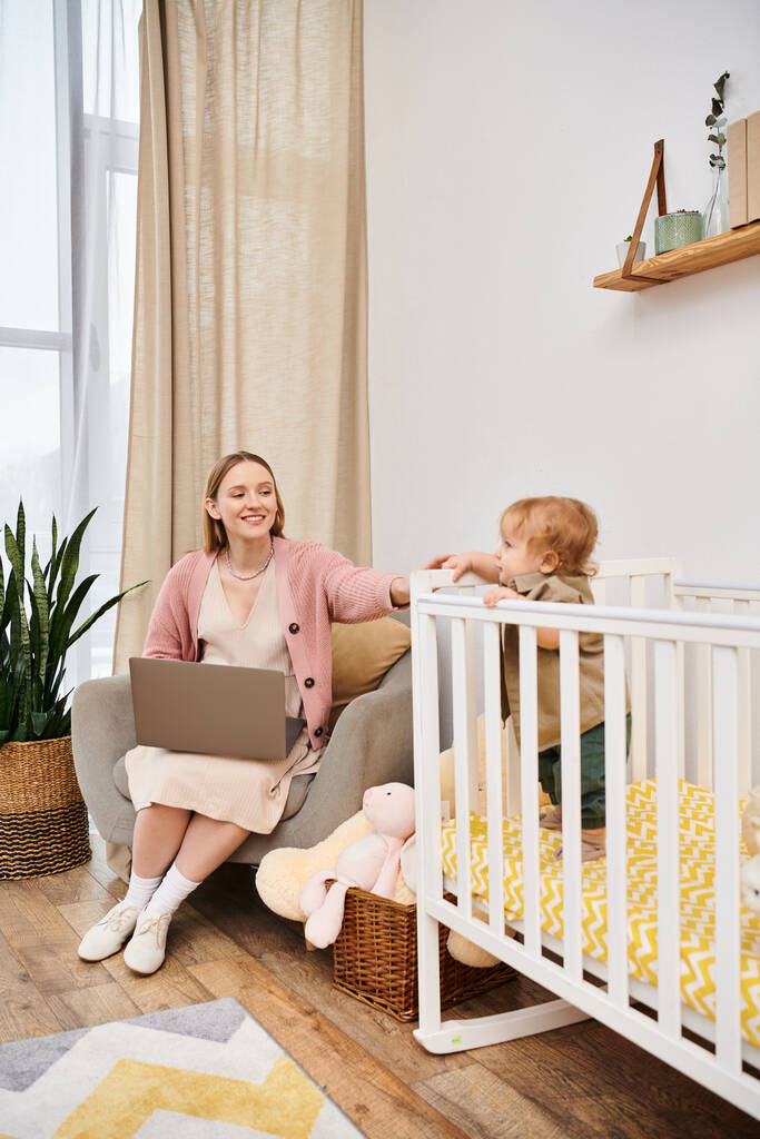 radosna kobieta pracująca na laptopie przy uroczym dziecku w łóżeczku niemowlęcym w pokoju dziecinnym, wielozadaniowa matka - Zdjęcie, obraz