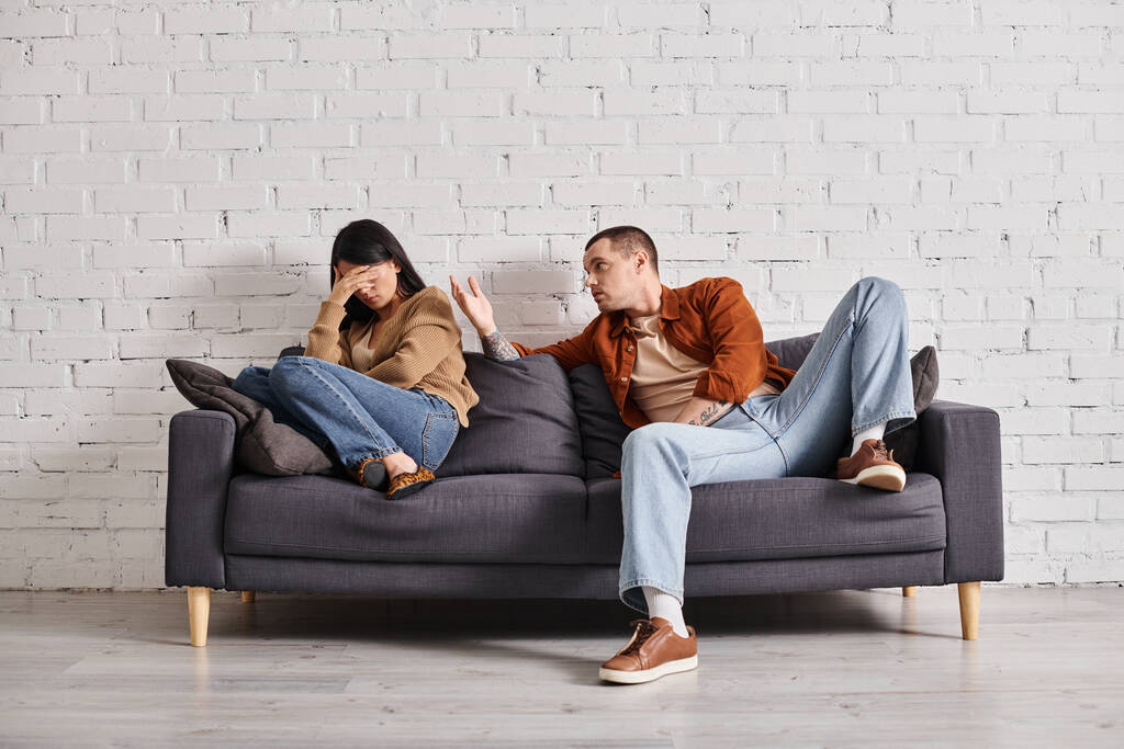 молодой эмоциональный мужчина разговаривает с азиаткой плачущей на диване в гостиной, концепция развода - Фото, изображение
