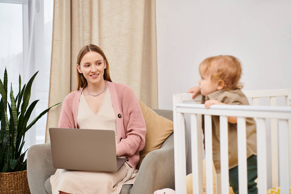 szczęśliwy matka pracuje na laptopie w pobliżu słodkie dziecko w łóżeczko dziecięce w pokoju dziecinnym, wielozadaniowa kobieta - Zdjęcie, obraz
