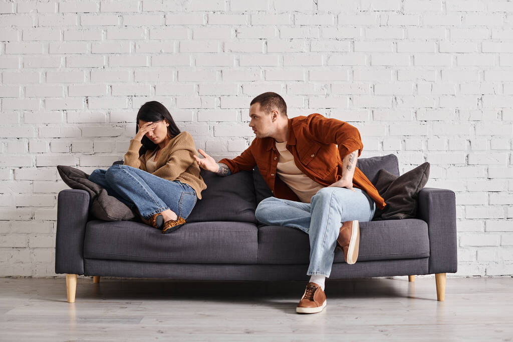 young irritated man talking to young asian wife crying on couch in living room, divorce concept - Photo, Image