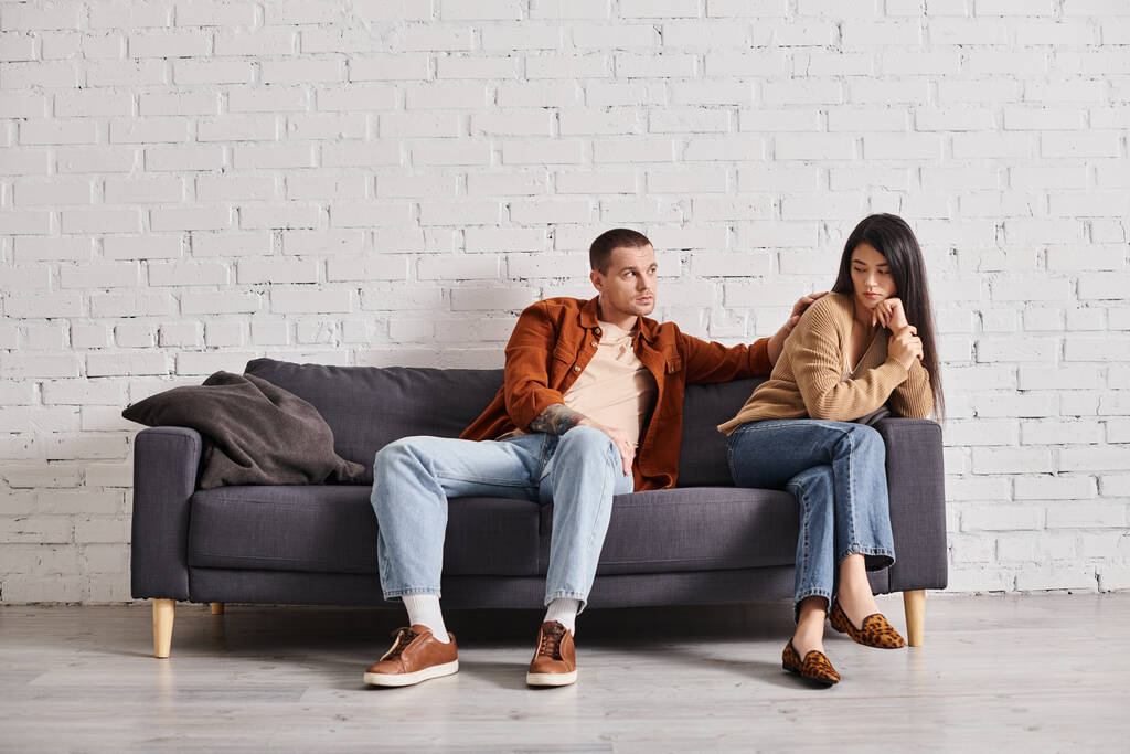 young man calming upset asian wife sitting on couch in living room, relationship difficulties - Photo, Image