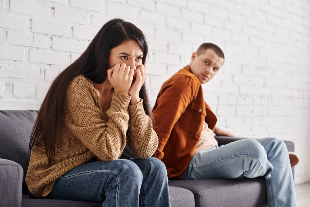 недовольный мужчина смотрит на обиженную азиатскую жену на диване в гостиной, отношения трудности - Фото, изображение