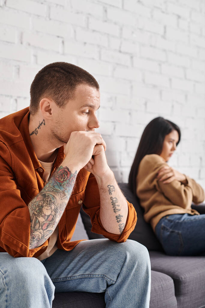 απογοητευμένος άντρας με τατουάζ που κοιτάζει προσβεβλημένη Ασιάτισσα σύζυγο που κάθεται στον καναπέ στο σπίτι, έννοια του διαζυγίου - Φωτογραφία, εικόνα