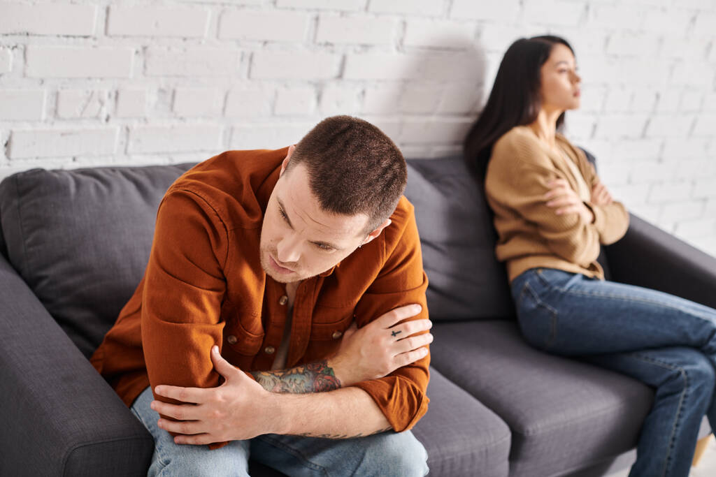 похмурий татуйований чоловік сидить на дивані у вітальні біля ображеної азіатської дружини, сімейна проблема - Фото, зображення