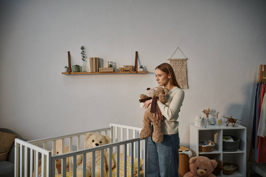Hilflose und aufgebrachte Frau mit Stofftier steht neben Krippe in trostlosem Kinderzimmer zu Hause - Foto, Bild