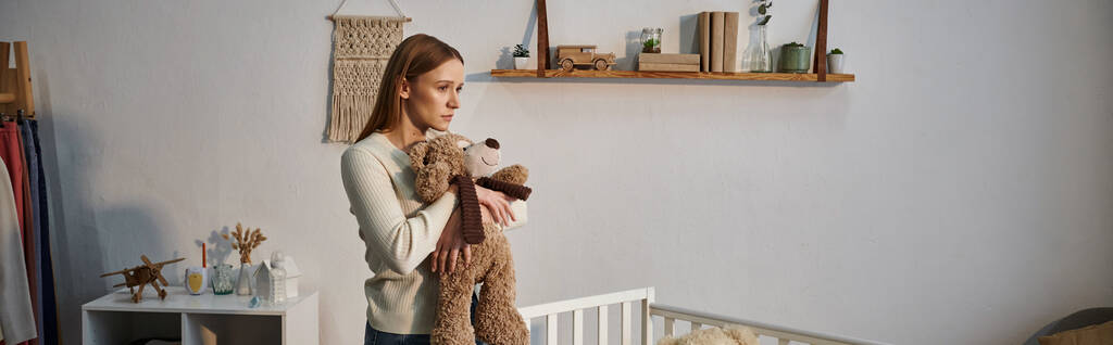 подавленная молодая женщина с мягкой игрушкой, стоящая возле кроватки в темной детской комнате дома, баннер - Фото, изображение