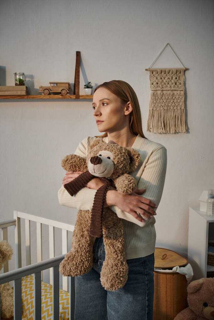 zware rouwende vrouw met zacht speelgoed staat in de buurt van wieg in sombere kinderkamer thuis - Foto, afbeelding