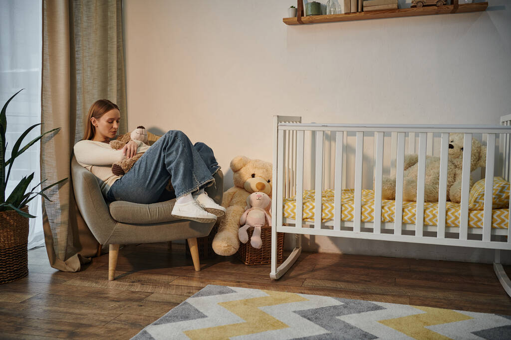 спустошена молода жінка з м'якою іграшкою, що сидить у кріслі біля дитячого ліжечка в похмурій кімнаті вдома - Фото, зображення
