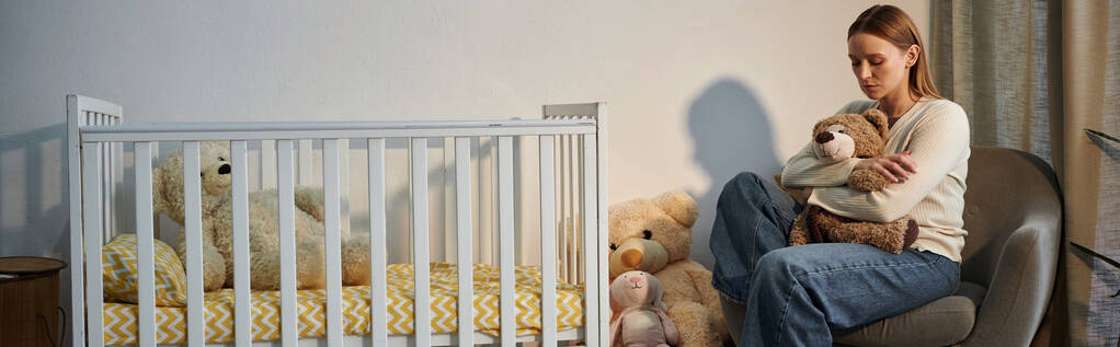 depressed young woman with soft toy sitting in armchair near crib in dark nursery room, banner - Photo, Image