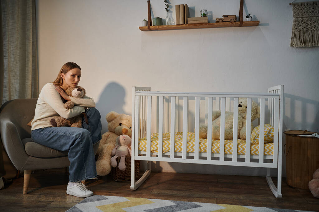 beznadějná mladá žena s měkkou hračkou sedí v křesle v blízkosti postýlky s v ponurém dětském pokoji doma - Fotografie, Obrázek