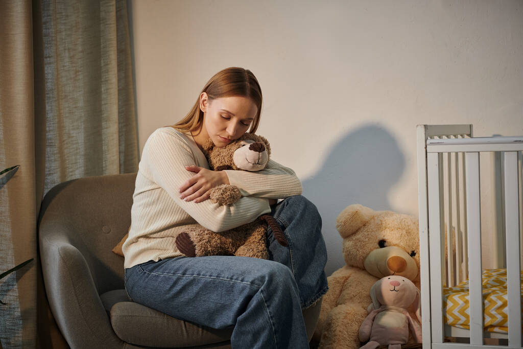 сумна сльозова жінка з м'якою іграшкою, що сидить у кріслі біля дитячого ліжечка в похмурій кімнаті вдома - Фото, зображення