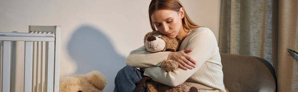 jovem deprimida com brinquedo macio sentado em poltrona perto berço em quarto berçário sombrio, banner - Foto, Imagem