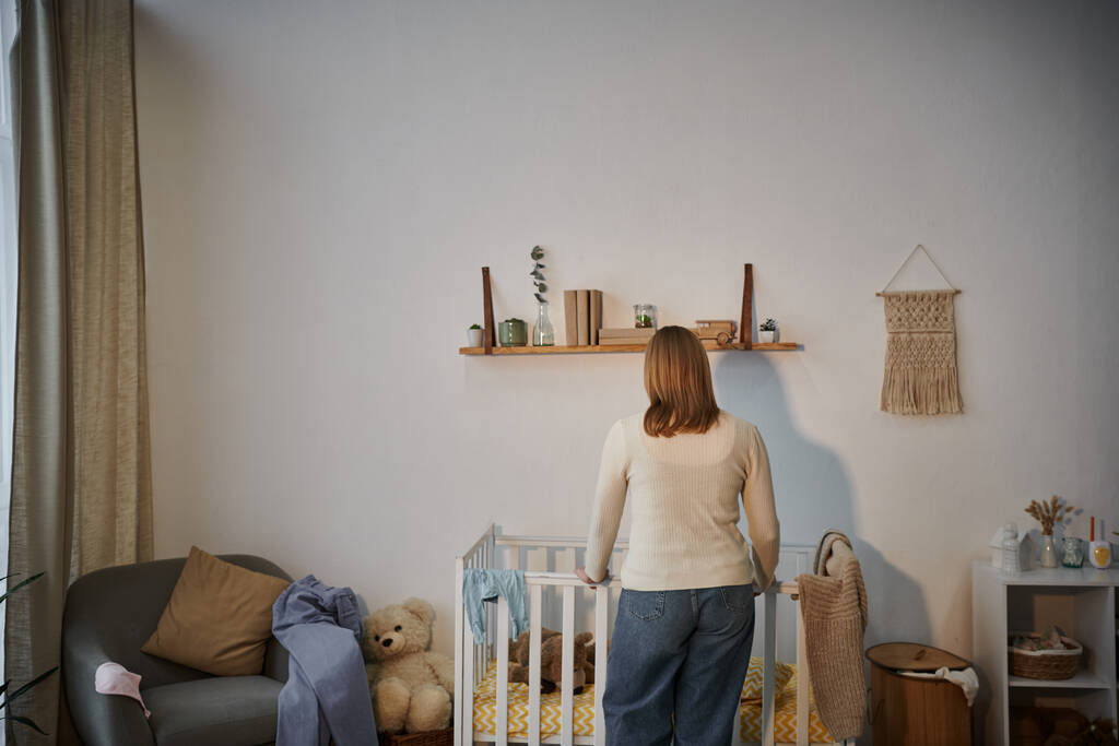 back view of depressed and lonely woman near crib with soft toys un dark nursery room at home - Photo, Image