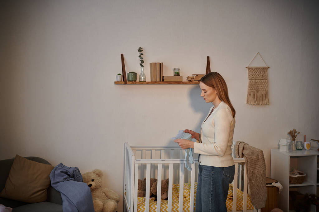 vista laterale della donna frustrata che tiene i vestiti per bambini vicino alla culla con giocattoli morbidi nella stanza dei bambini - Foto, immagini