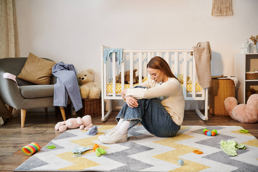 Junge unterlegene Frau sitzt mit gesenktem Kopf neben Spielzeug und Krippe im Kinderzimmer zu Hause - Foto, Bild