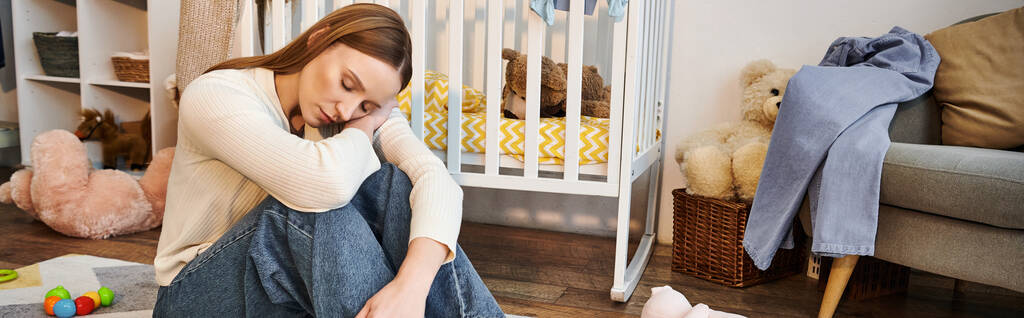 Kederli genç bir kadın, bebek odasındaki bebek karyolasının yanında gözleri kapalı, yerde oturuyor. - Fotoğraf, Görsel