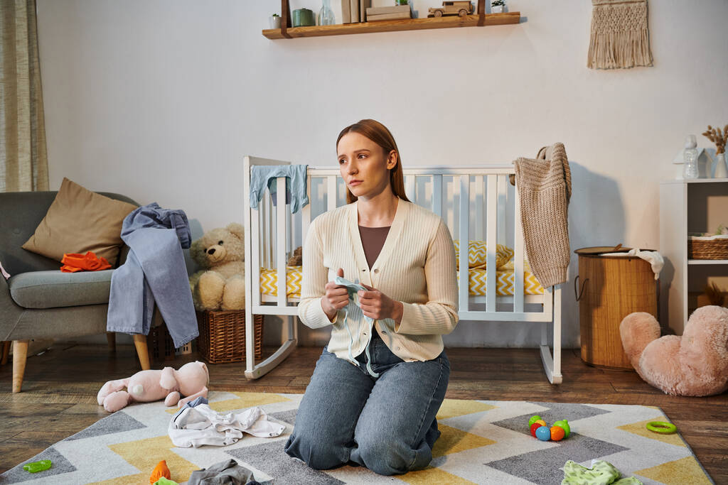 беспокойная женщина с детской одеждой сидит на полу рядом с кроваткой и игрушками в детской комнате дома - Фото, изображение
