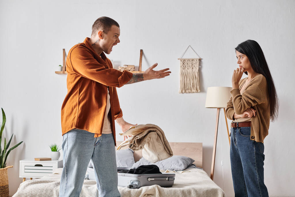 młody i wściekły mężczyzna kłócący się o azjatycką żonę w pobliżu walizki i rzeczy w sypialni w domu - Zdjęcie, obraz
