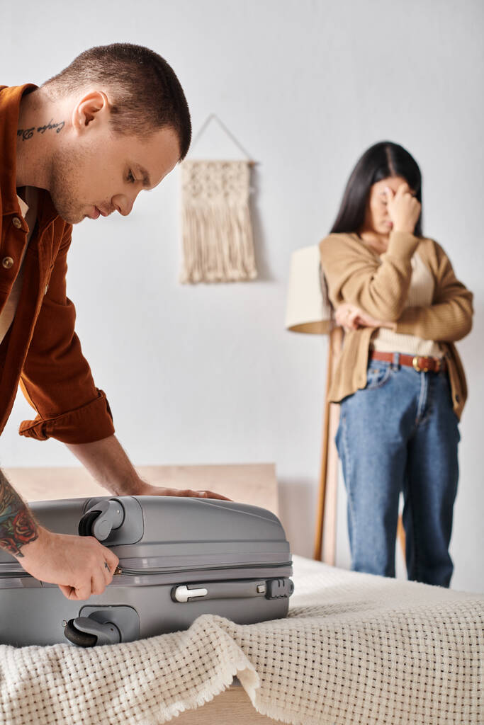 young man packing suitcase near depressed asian woman crying in bedroom at home, family divorce - Photo, Image