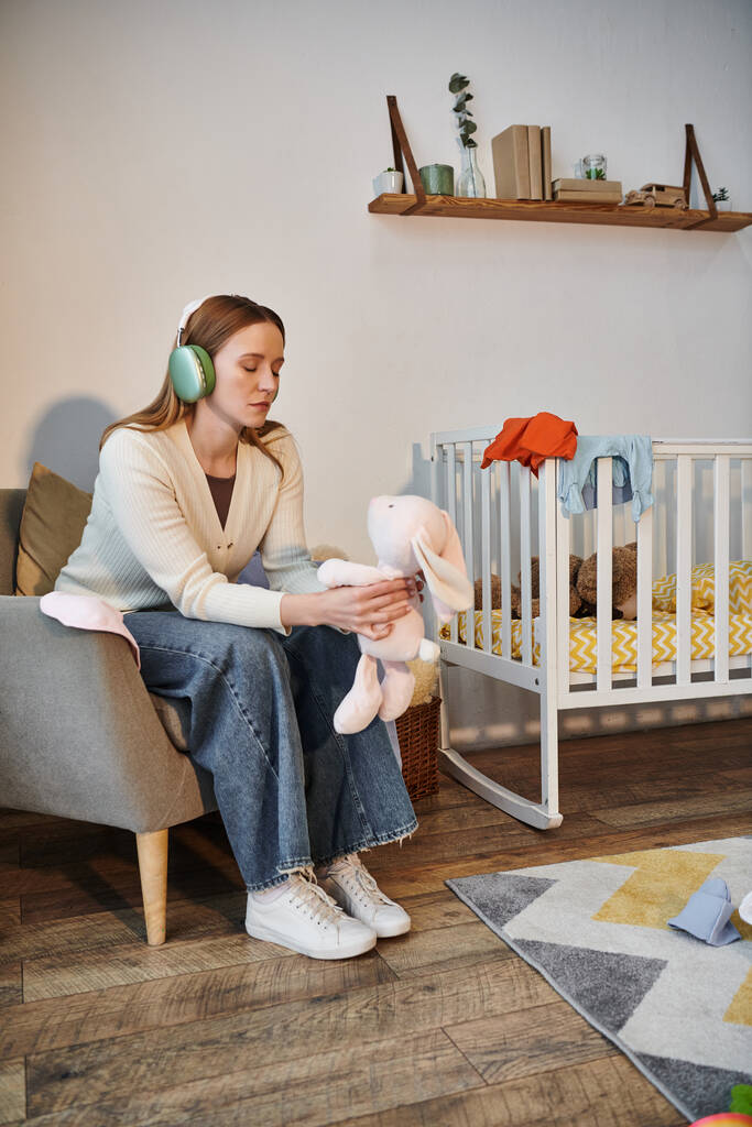 απελπισμένη γυναίκα κρατώντας μαλακό παιχνίδι και ακούγοντας μουσική στα ακουστικά στο σκοτεινό παιδικό δωμάτιο στο σπίτι - Φωτογραφία, εικόνα
