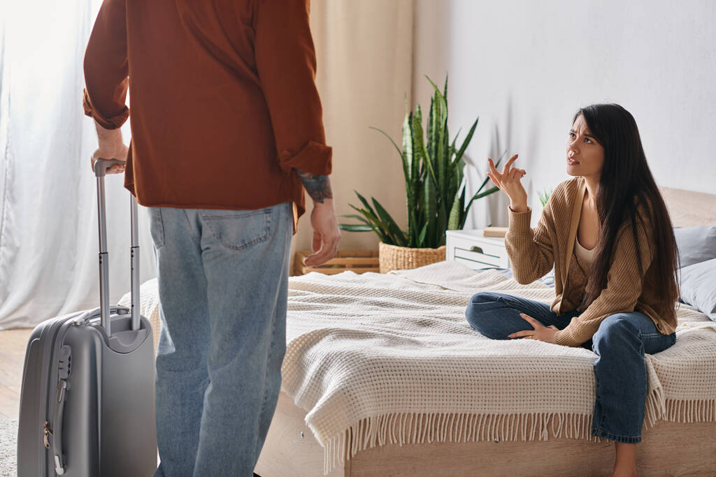 boos aziatische vrouw zitten op bed en praten op echtgenoot met koffer thuis, echtscheiding concept - Foto, afbeelding