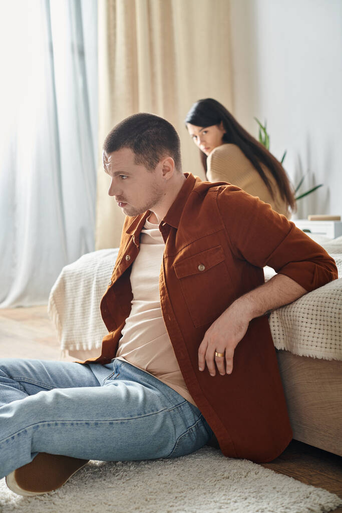 fiatal sértett férfi ül padlón közelében ideges ázsiai feleség, kapcsolat nehézségek koncepció - Fotó, kép