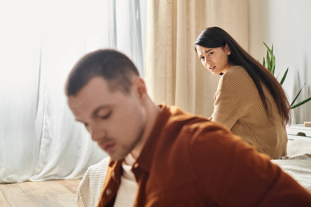δυσαρεστημένοι συνοφρυωμένη Ασιάτισσα γυναίκα κοιτάζοντας τον νεαρό αναστατωμένο σύζυγο στο υπνοδωμάτιο στο σπίτι, έννοια του διαζυγίου - Φωτογραφία, εικόνα
