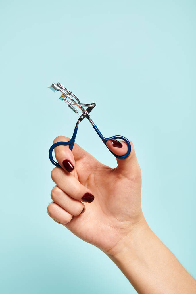 objeto foto de rizador de pestañas en la mano de mujer joven desconocida con esmalte de uñas sobre fondo azul - Foto, Imagen