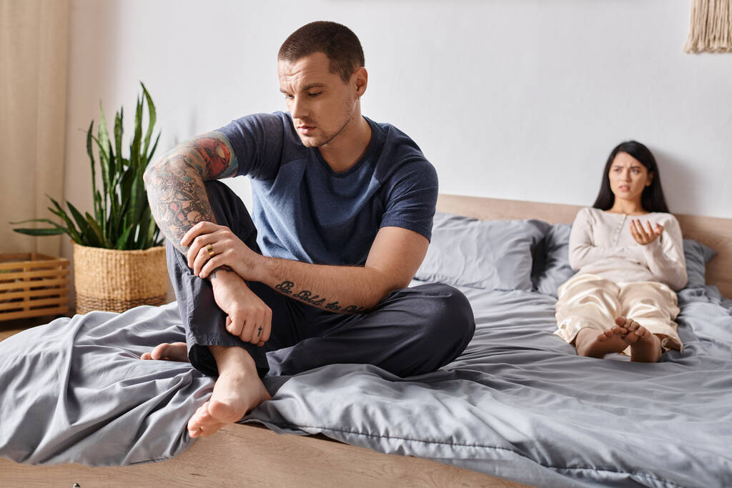 zdenerwowany wytatuowany mężczyzna siedzi na łóżku blisko sfrustrowany azjatycki żona, stosunki rodzinne trudności - Zdjęcie, obraz