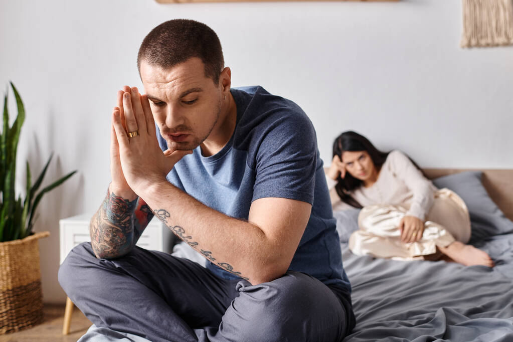νεαρός καταθλιπτικός άνδρας με τατουάζ που κάθεται στο κρεβάτι κοντά στην προσβεβλημένη Ασιάτισσα σύζυγο, έννοια του οικογενειακού διαζυγίου - Φωτογραφία, εικόνα