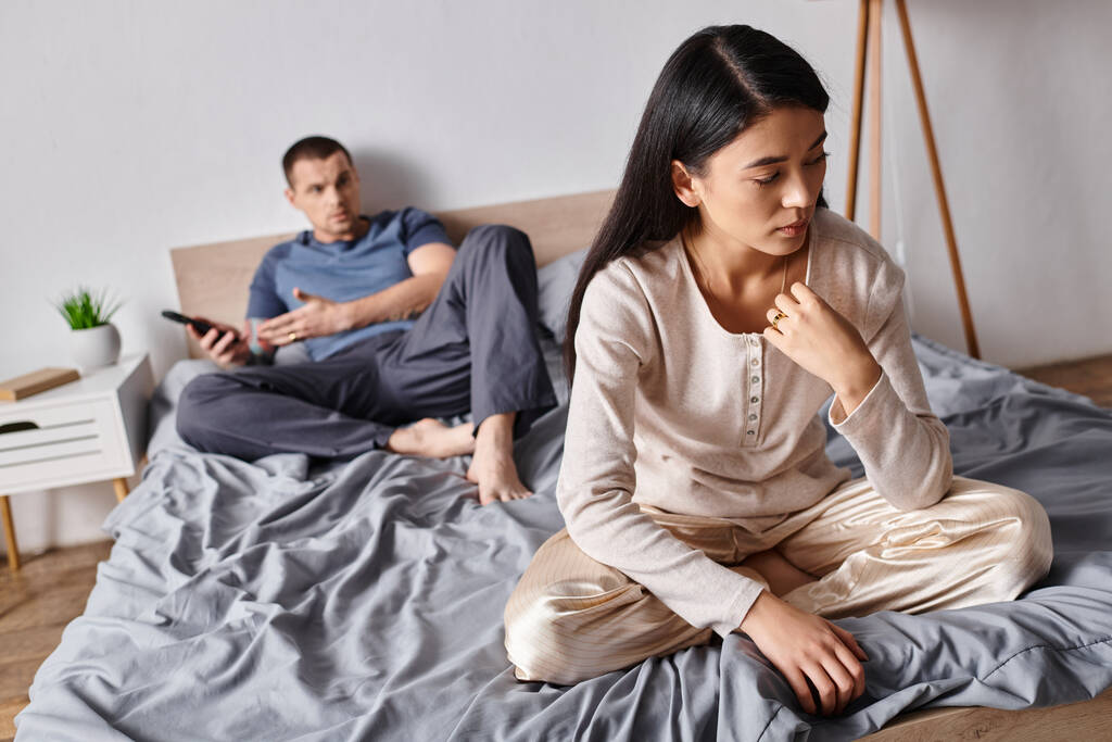 απογοητευμένη Ασιάτισσα γυναίκα κάθεται κοντά στο σύζυγο χρησιμοποιώντας smartphone στο υπνοδωμάτιο στο σπίτι, οικογενειακό πρόβλημα - Φωτογραφία, εικόνα