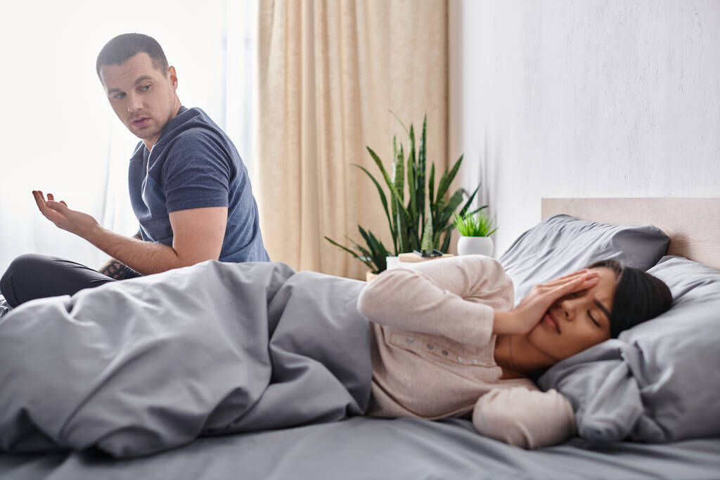 イライラするアジアの妻に話す若い男 自宅でベッドに顔を隠す, 関係の困難 - 写真・画像