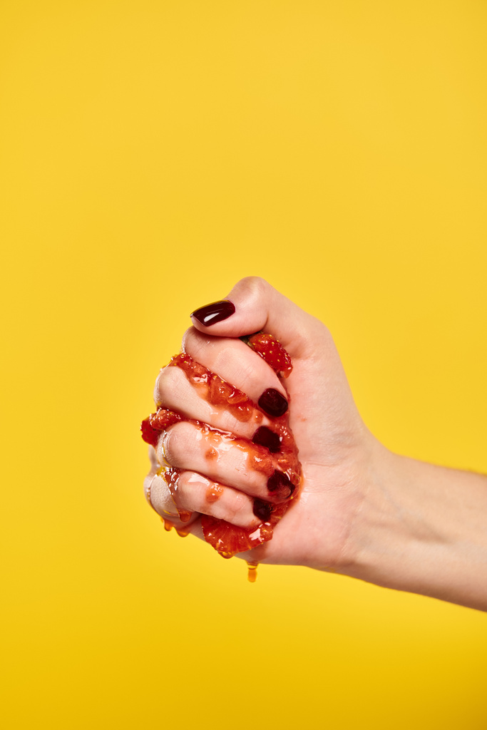 άγνωστο νεαρό θηλυκό μοντέλο που συμπιέζει κόκκινες ζουμερές φράουλες στο χέρι της σε κίτρινο φόντο - Φωτογραφία, εικόνα