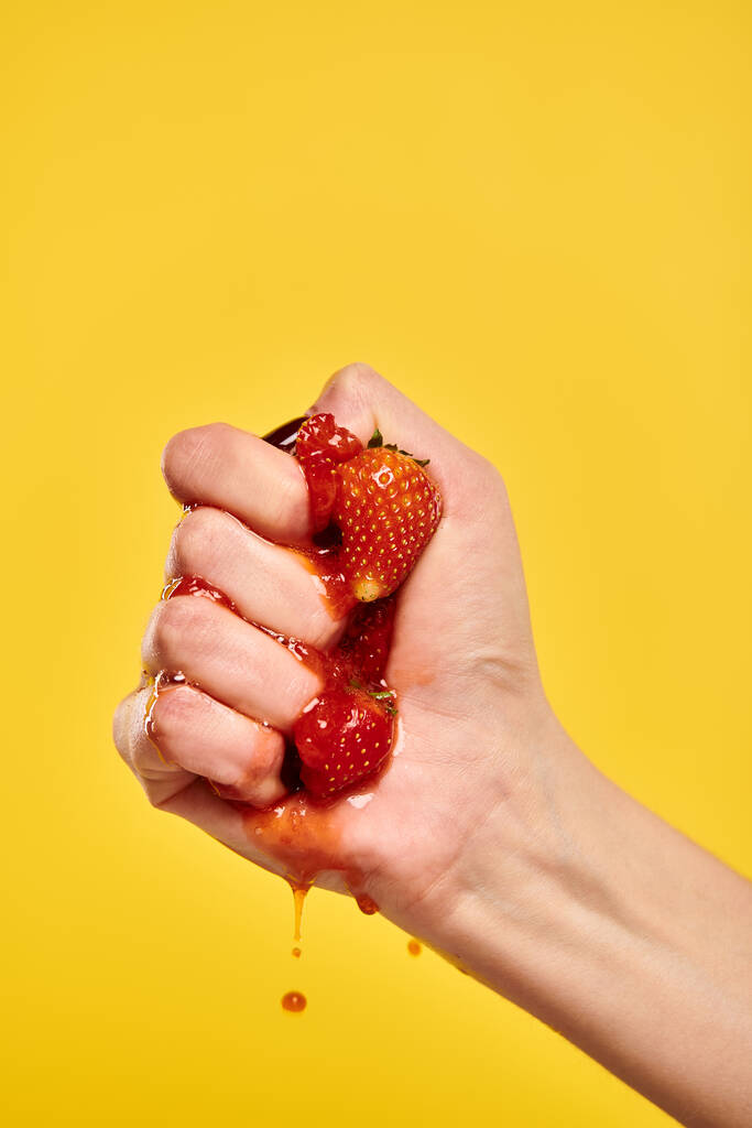 unknown young woman with nail polish squeezing red juicy fresh strawberries on yellow background - Photo, Image