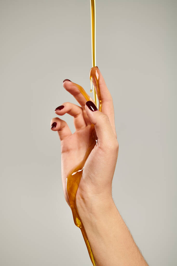 ręka nieznanej młodej kobiety z lakierem do paznokci pokrytym słodkim świeżym miodem na szarym tle - Zdjęcie, obraz