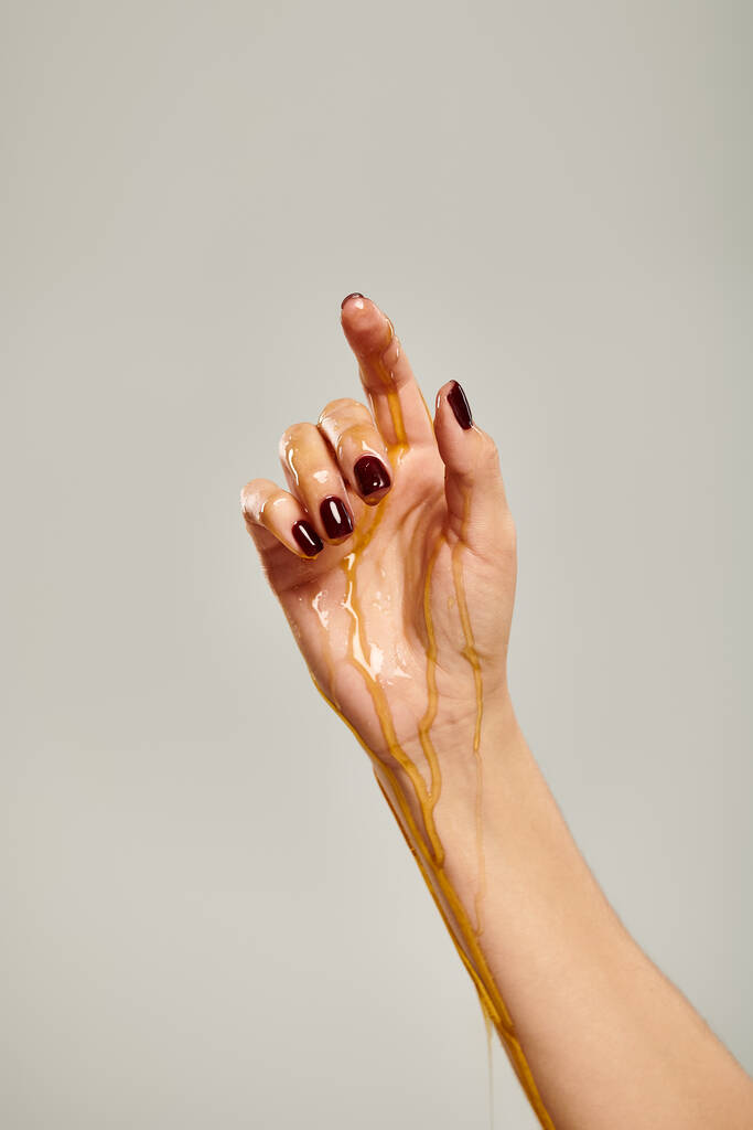 mano de modelo femenino desconocido con esmalte de uñas cubierto con miel deliciosa orgánica sobre fondo gris - Foto, imagen