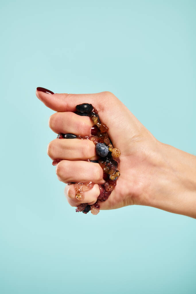 невідома молода жінка з лаком для нігтів стискає свіжу соковиту чорницю в руці на синьому фоні - Фото, зображення