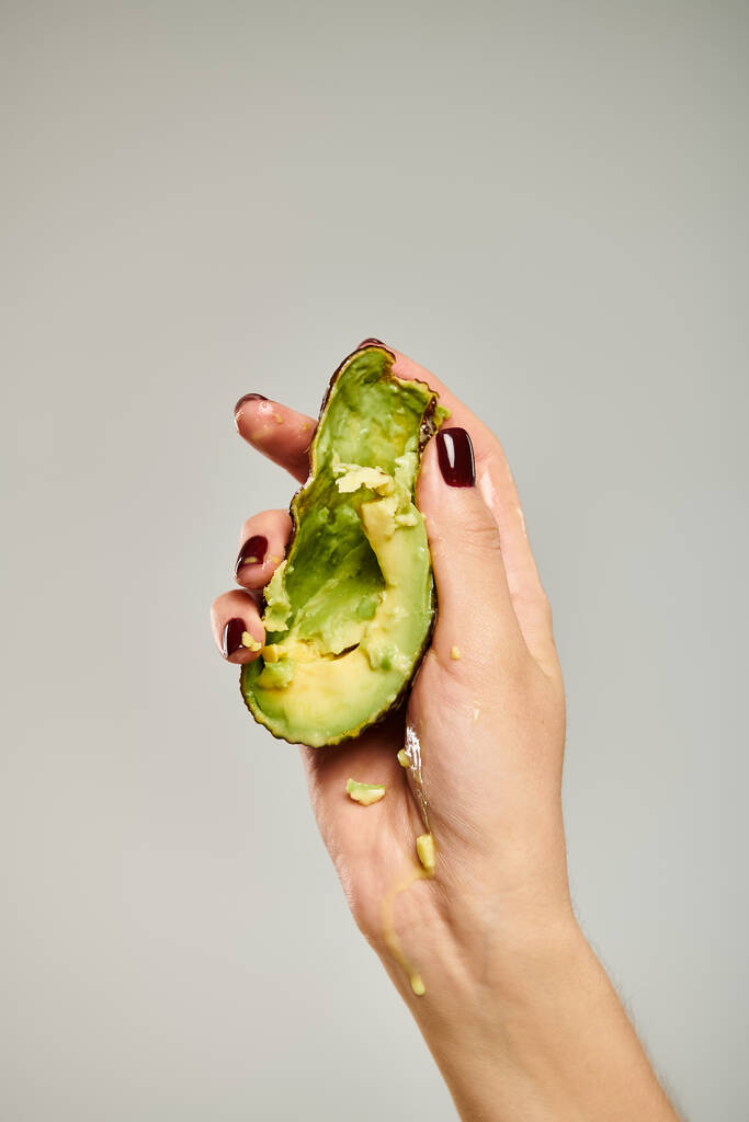Unbekanntes weibliches Modell drückt gesunde köstliche Avocado in die Hand, während sie auf grauem Hintergrund sitzt - Foto, Bild