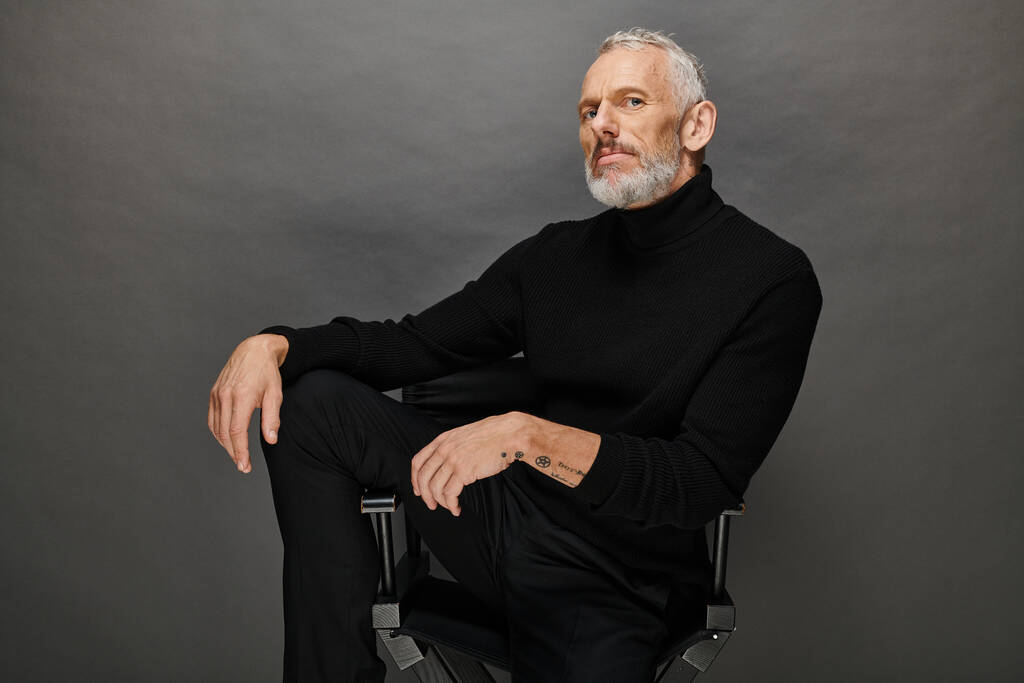 appealing mature man with beard in voguish turtleneck sitting on folding chair and looking at camera - Photo, Image